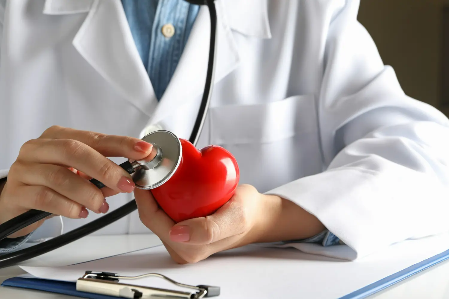 Médico usa um estetoscópio para avaliar a miniatura de um coração, simbolizando os exames do coração